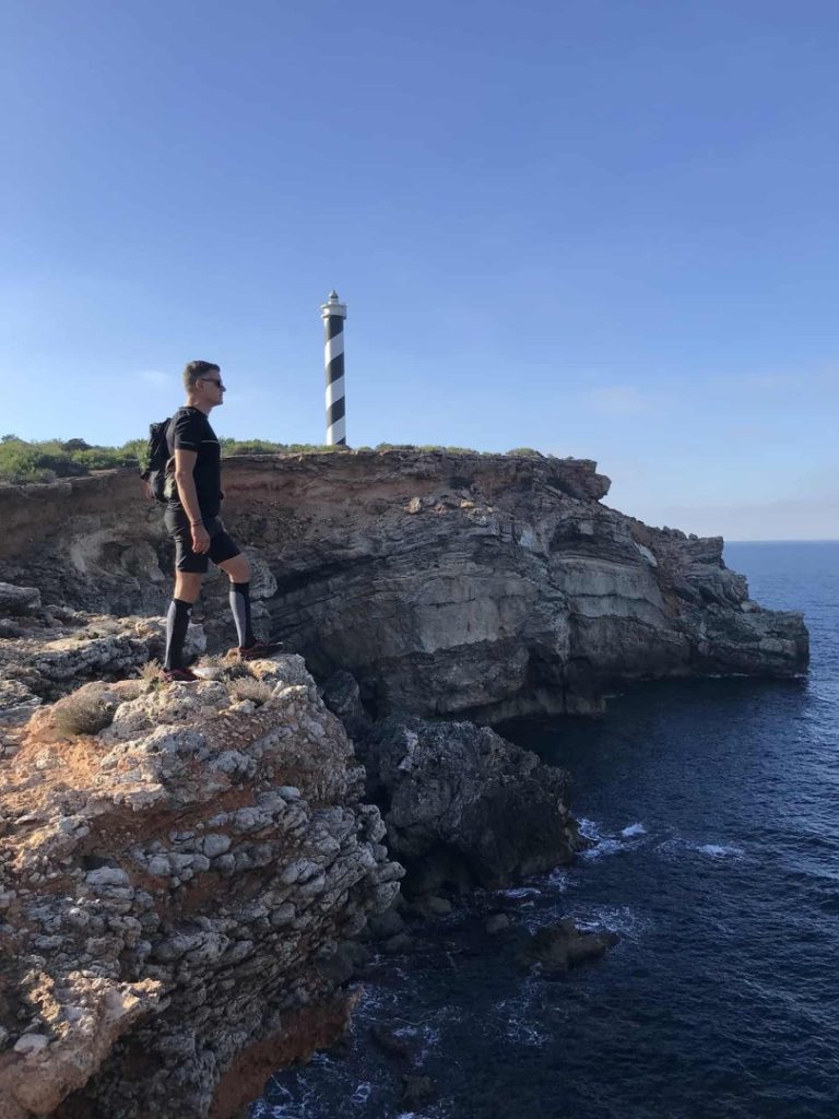 Hiker on coastal cliff near striped lighthouse during Azure Hike Station scenic coastal hike in Ibiza