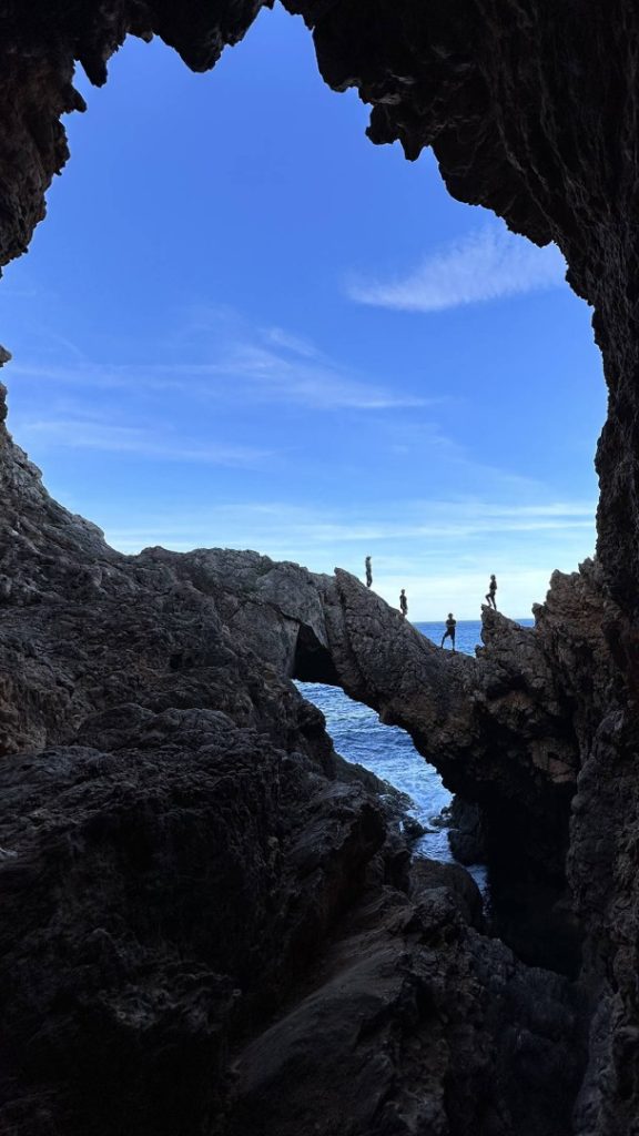 Hikers crossing a natural rock bridge inside a sea cave on Ibiza’s memorable Deus Hike Station coastal trail