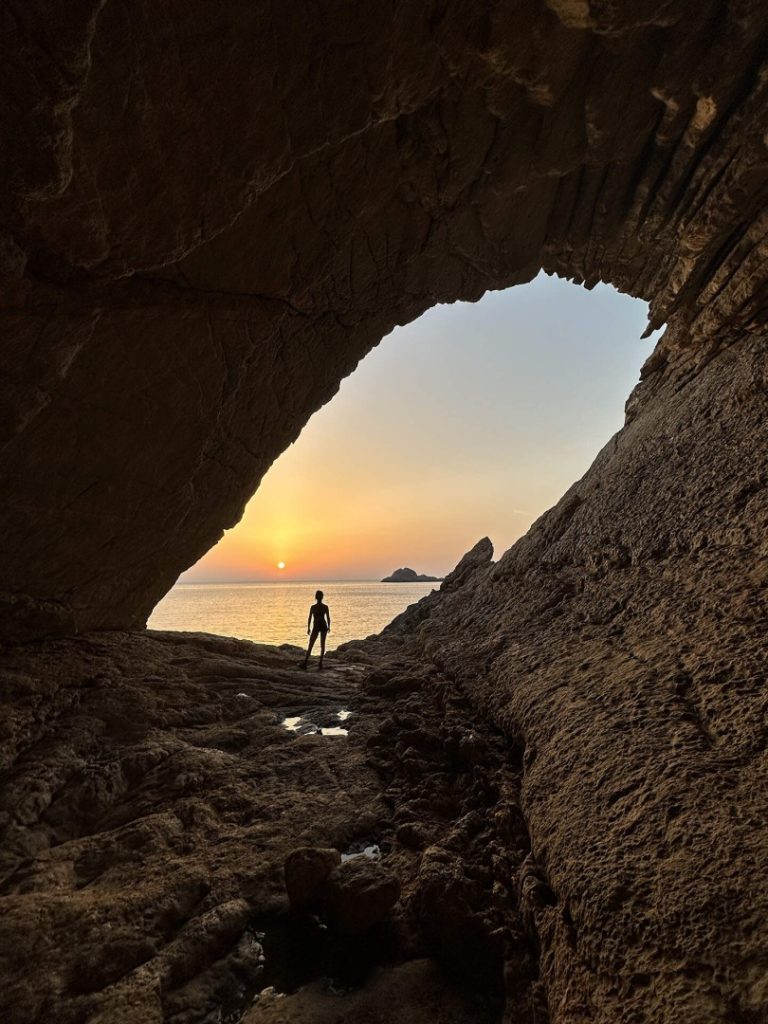 Hiker silhouetted in a sea cave at sunset enjoying the view on Ibiza’s dramatic Deus Hike Station coastal trail