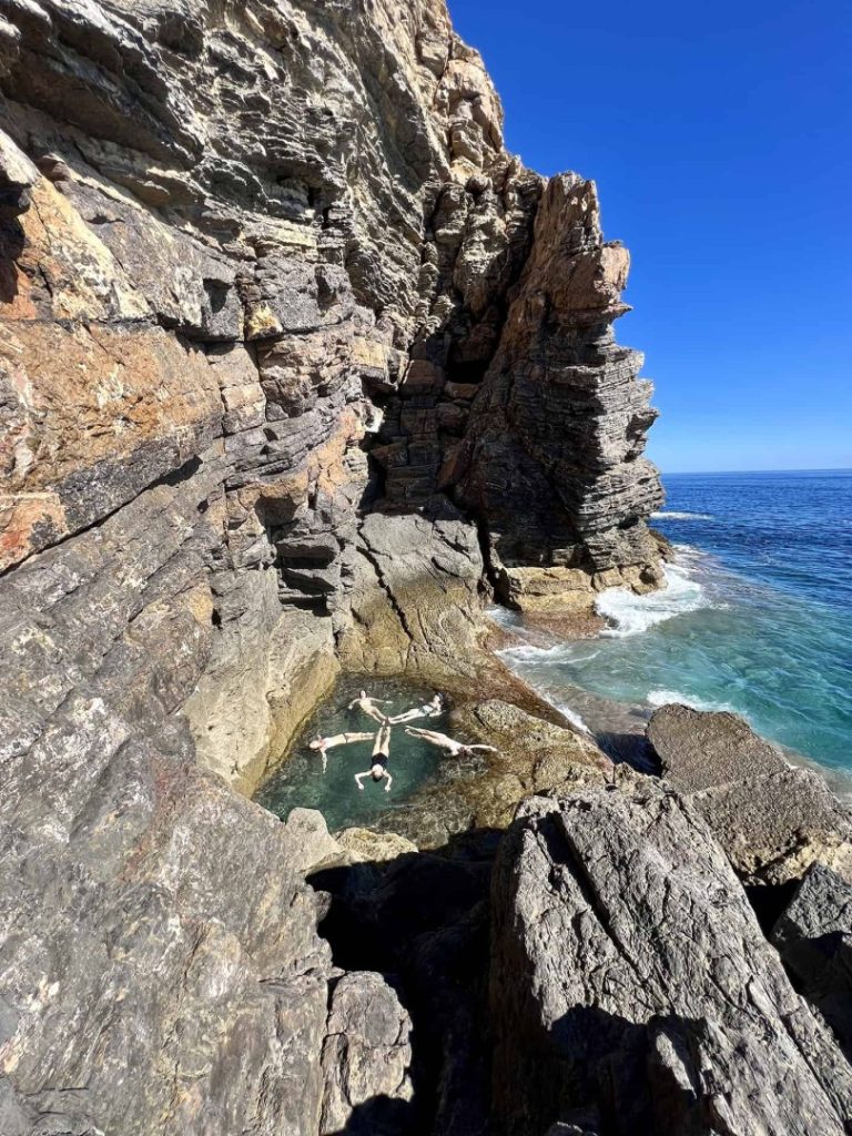 Hikers relaxing in a natural rock pool beside impressive cliffs and turquoise sea on Ibiza’s Stone Hike Station coastal trail