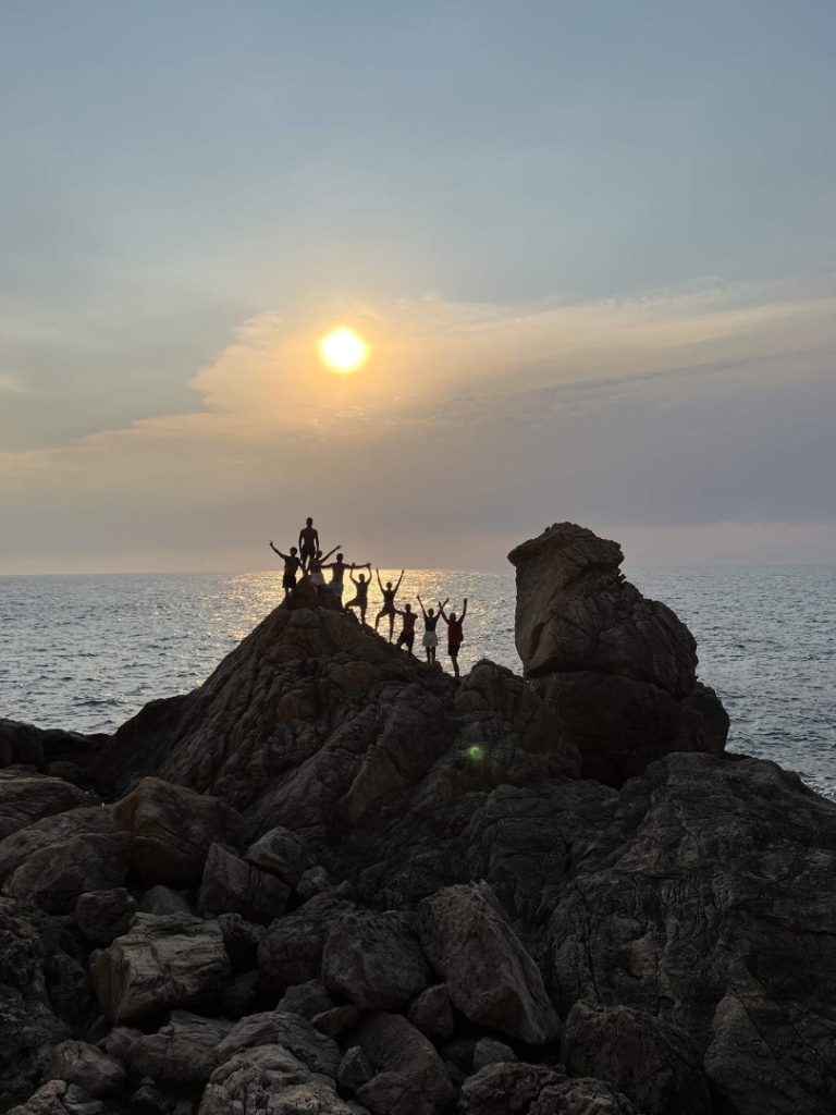 Hiking group celebrating on coastal rocks at sunset above the Mediterranean on Ibiza’s scenic Lagoon Hike Station trail