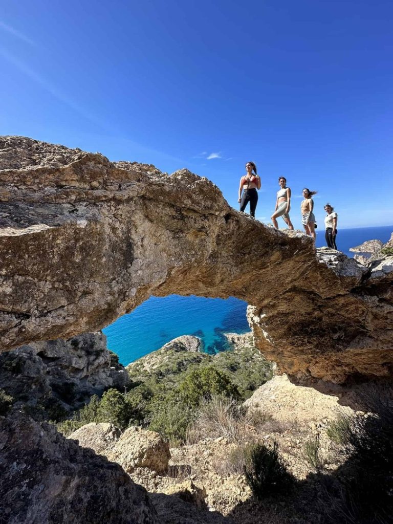 Hikers posing for a picture on a natural rock arch above turquoise Mediterranean waters on Ibiza’s scenic Legend Hike Station trail