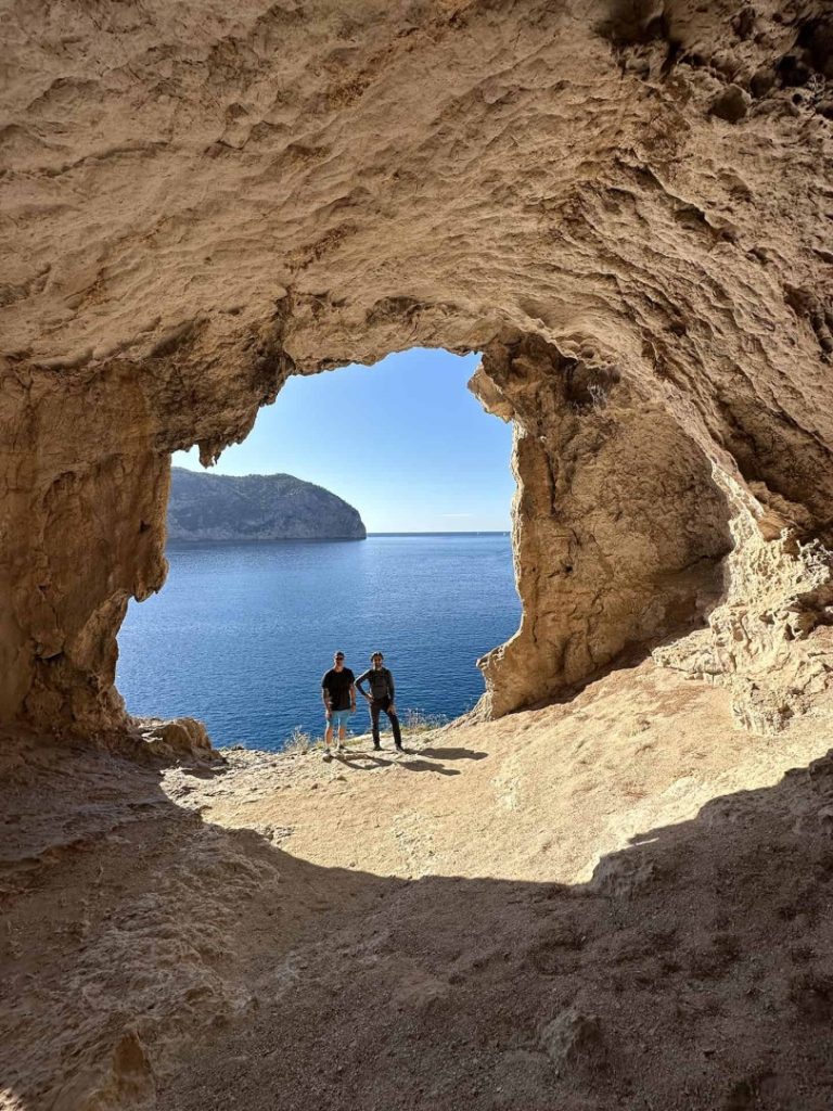 Hikers standing in natural sea cave arch overlooking Mediterranean coast on Quelona Hike Station coastal hike in Ibiza
