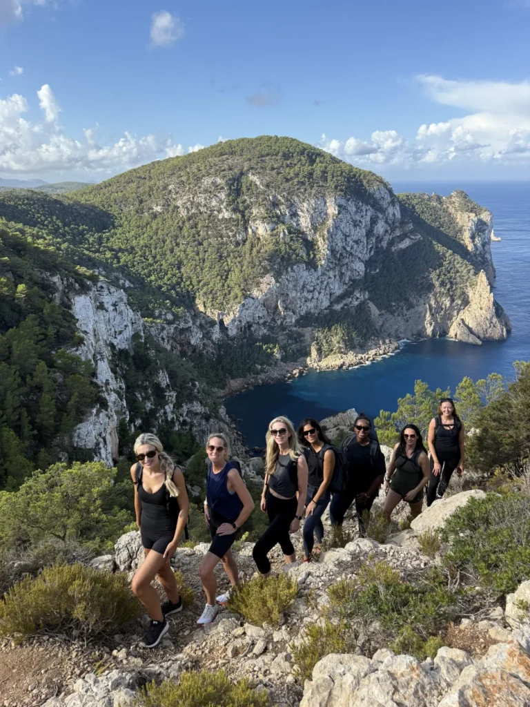 Group hiking above Cala d’Albarca cliffs on Ibiza’s North Hike Station trail with panoramic Mediterranean cove view.